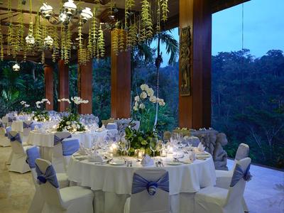 Elegant dinner setup with round tables, white chairs, and flower decorations near large glass windows.