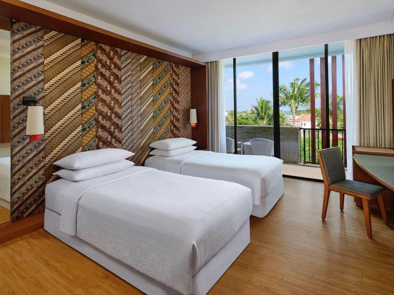 Jasny pokój hotelowy z dwoma pojedynczymi łóżkami, drewnianą dekoracją ściany i balkonem z widokiem na palmy.