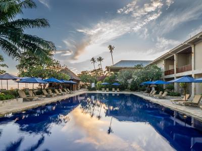 Une piscine d'hôtel avec des parasols bleus, des palmiers et un ciel clair avec des nuages au coucher du soleil.