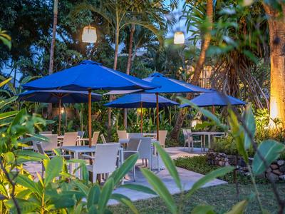 Terrasse de jardin en soirée avec des tables, des chaises et des parasols bleus dans un cadre tropical.
