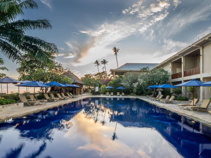 Large outdoor pool with palm trees and blue umbrellas beside a hotel at sunset.