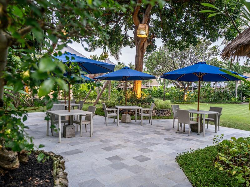 Outdoor seating area with tables, chairs, and blue umbrellas on stone pavement surrounded by greenery.