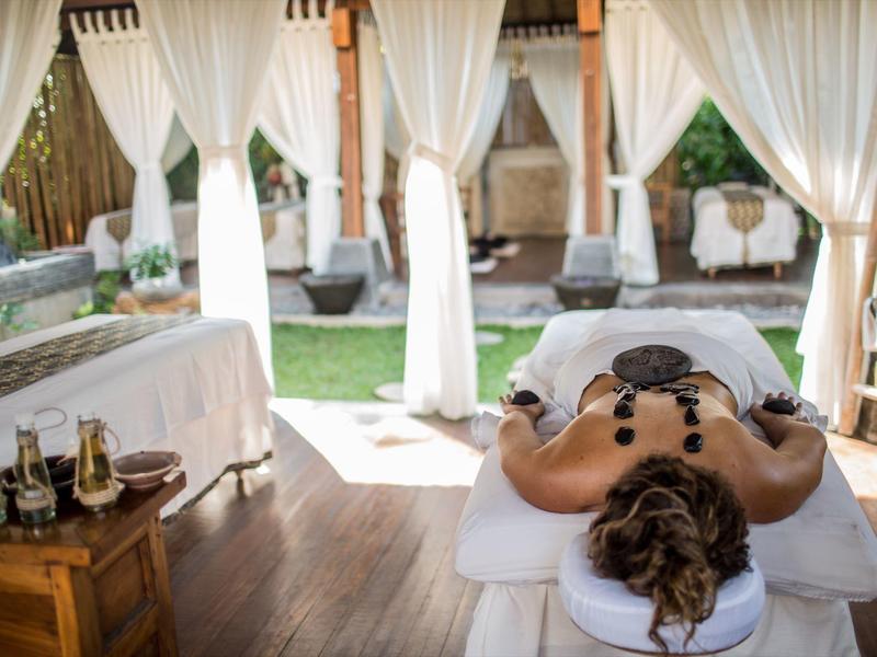 Person receiving hot stone massage in an open spa area with white curtains