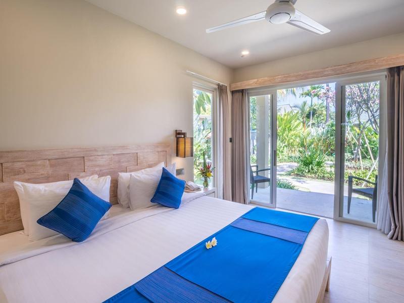 Bright hotel room with two beds, blue accents, ceiling fan, and view of tropical garden.