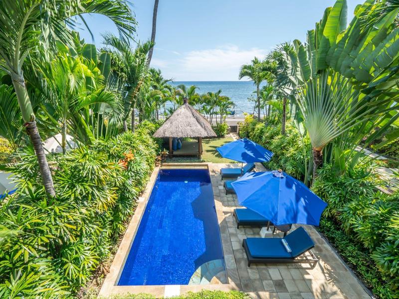 A luxury pool area with blue umbrellas and palm trees, overlooking the sea.
