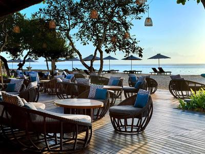 Strandrestaurant mit Holzboden, runden Tischen, Rattanstühlen und Blick auf Meer mit Sonnenschirmen.