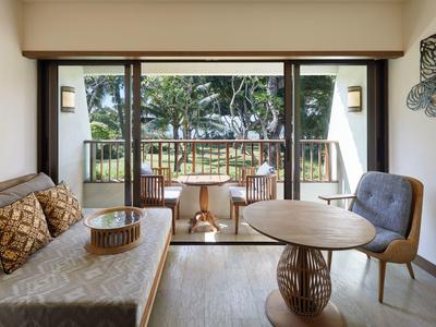 Chambre d'hôtel lumineuse avec canapé, chaise, table et balcon avec vue sur la verdure.
