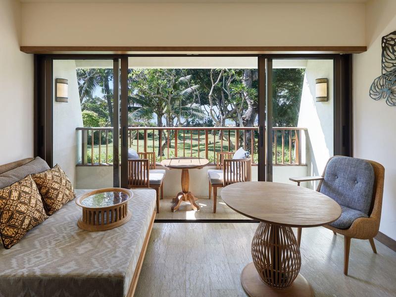 Chambre d'hôtel lumineuse avec canapé, chaise, table et balcon avec vue sur la verdure.