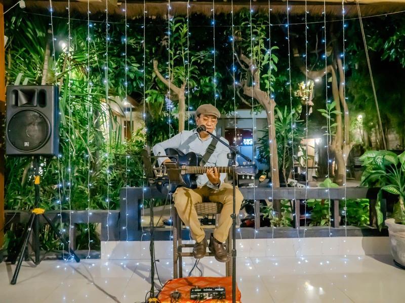 Mężczyzna siedzi z gitarą przed zieloną oświetloną ścianą ze zwisającymi światełkami.