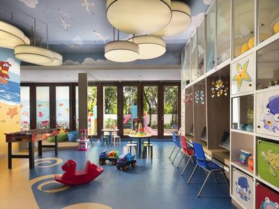 Sala de juegos infantil con sillas coloridas, juguetes y grandes lámparas bajo un cielo azul pintado en el techo.