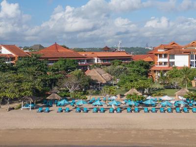 Spiaggia con lettini azzurri e ombrelloni davanti a un complesso alberghiero con tetti rossi.