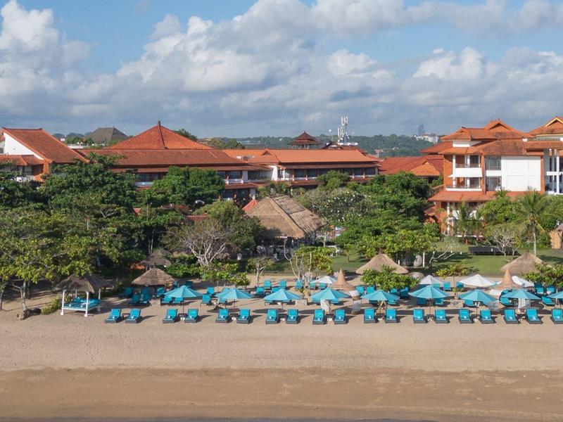 Spiaggia con lettini azzurri e ombrelloni davanti a un complesso alberghiero con tetti rossi.