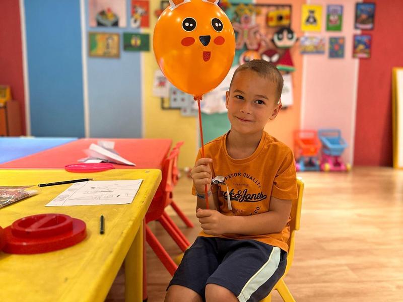 Bambino seduto che tiene un palloncino arancione a forma di faccia in una stanza colorata.