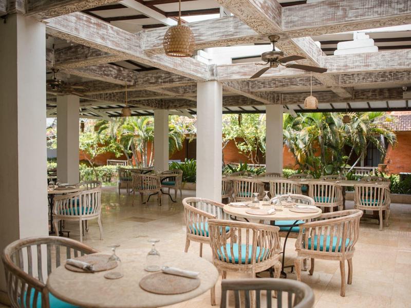 Terrazza ombreggiata con tavoli rotondi e sedie in legno, atmosfera tropicale e luminosa.