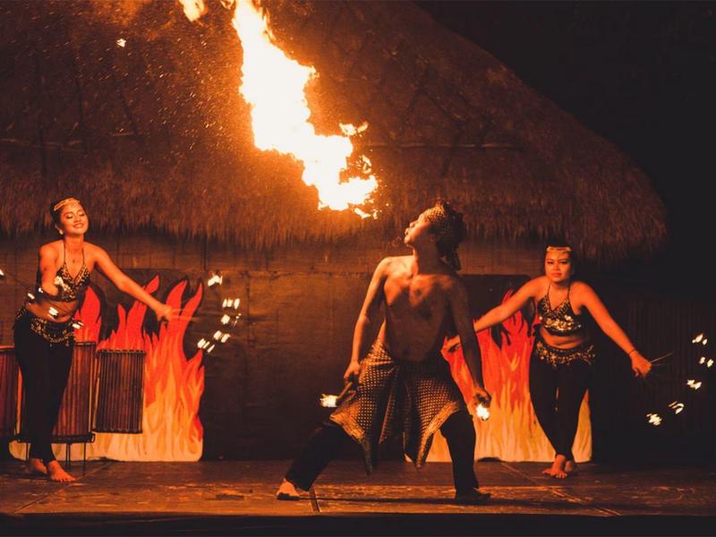 Spettacolo di danzatori tradizionali con fuoco su un palco scuro sotto un tetto di paglia.