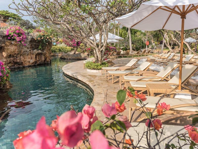 Espace piscine avec chaises longues et parasols, entouré de plantes et d'arbres en fleurs.