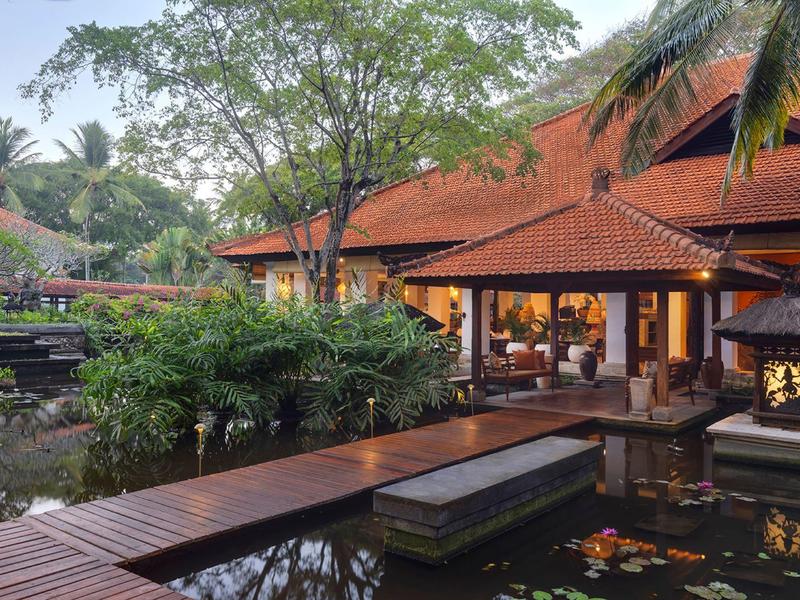 Hôtel à l'architecture traditionnelle entouré d'une végétation luxuriante et d'étangs avec passerelles en bois.