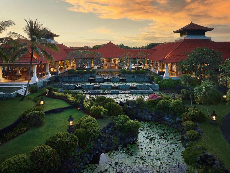 Vue en soirée d'une station tropicale avec piscine, jardins luxuriants et toits rouges.