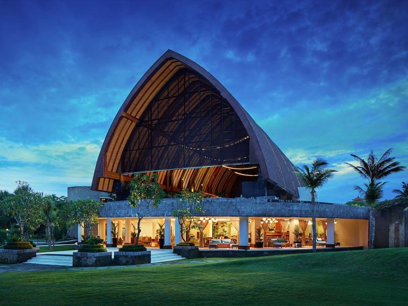 Nowoczesny budynek z dużym zakrzywionym dachem, oświetlony o zmierzchu, otoczony bujną zielenią.