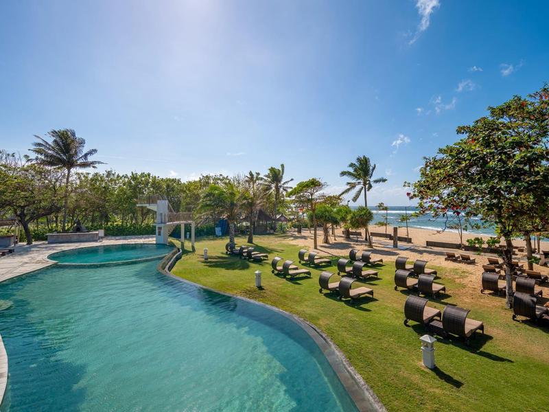 Tropikalny kurort z zakrzywionym basenem, leżakami, palmami i piaszczystą plażą pod niebieskim niebem.