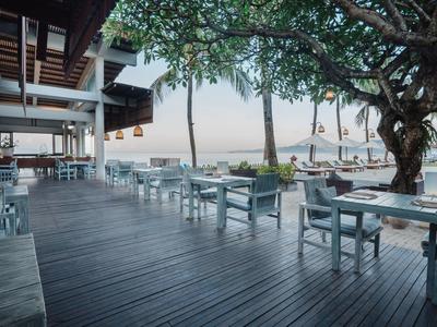 Terraza abierta de restaurante junto al mar con suelo de madera, mesas y sillas blancas bajo un árbol.