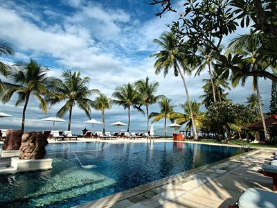 Una piscina al aire libre con tumbonas y palmeras frente a una playa de arena y cielo azul.
