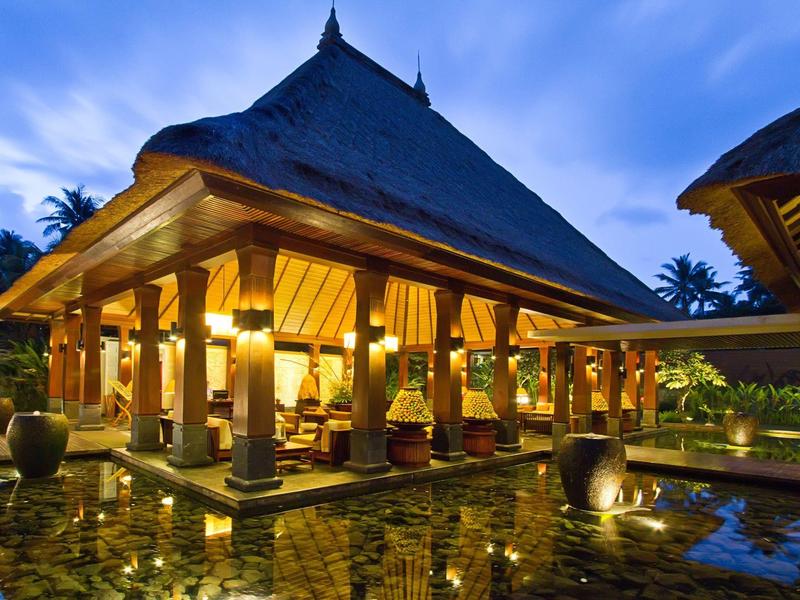 Pabellón iluminado con techo tradicional y reflejo en agua al atardecer en un resort tropical.