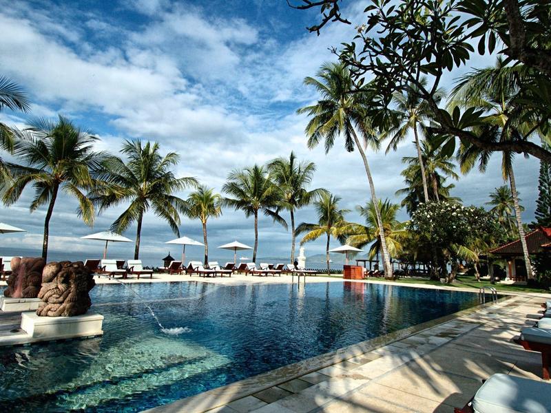 Una piscina al aire libre con tumbonas y palmeras frente a una playa de arena y cielo azul.