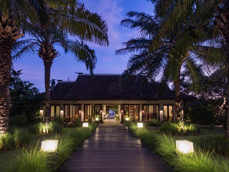 Un camino de jardín iluminado conduce a un hotel tropical con palmeras al anochecer.