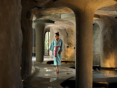 Eine Frau in blauem Kleid geht in einem dunklen, tropfsteinähnlichen Gang mit Säulen aus Stein.