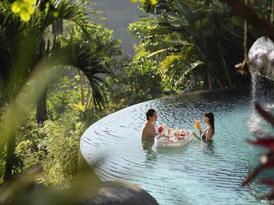 Zwei Menschen entspannen im Infinity-Pool, umgeben von üppigem, grünem Dschungel und Sonnenlicht.