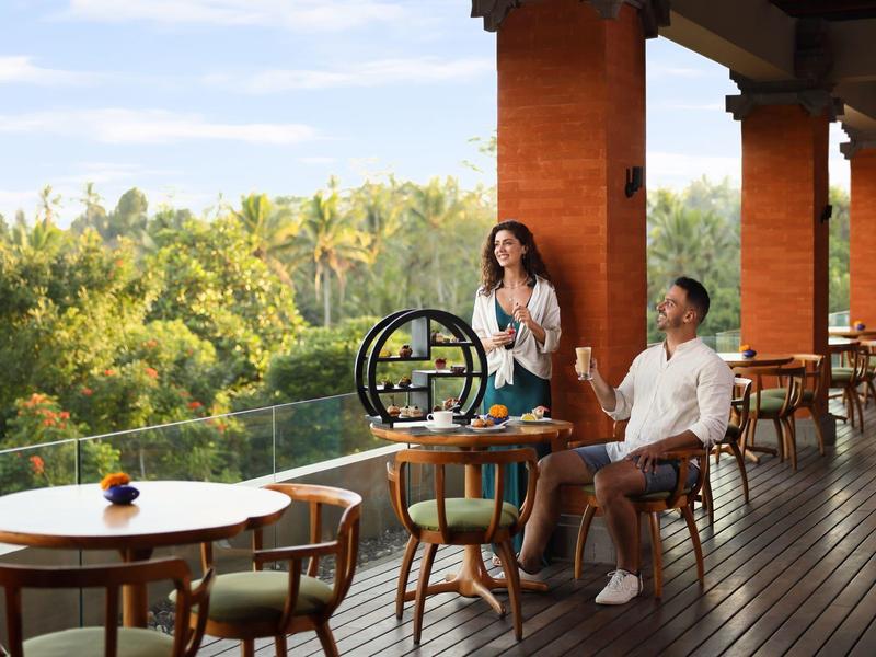 Offene Terrasse mit Holzboden, Tischen und Stühlen, umgeben von grüner Vegetation und blauem Himmel.