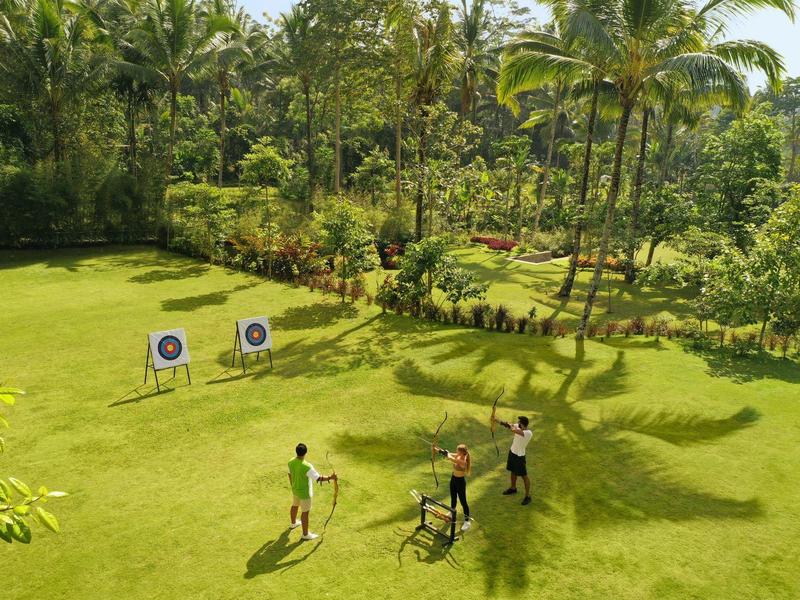 Drei Personen üben Bogenschießen auf grünem Rasen mit zwei Zielscheiben und Palmen im Hintergrund.