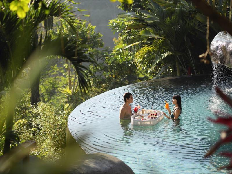 Zwei Menschen entspannen im Infinity-Pool, umgeben von üppigem, grünem Dschungel und Sonnenlicht.