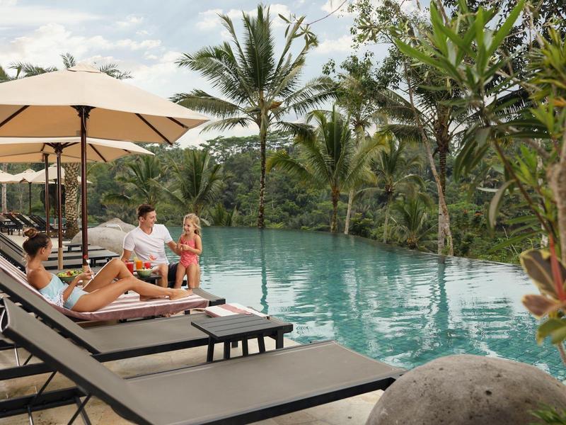 Pool mit Liegestühlen, Sonnenschirmen und Palmen, umgeben von grüner Landschaft bei bewölktem Himmel.