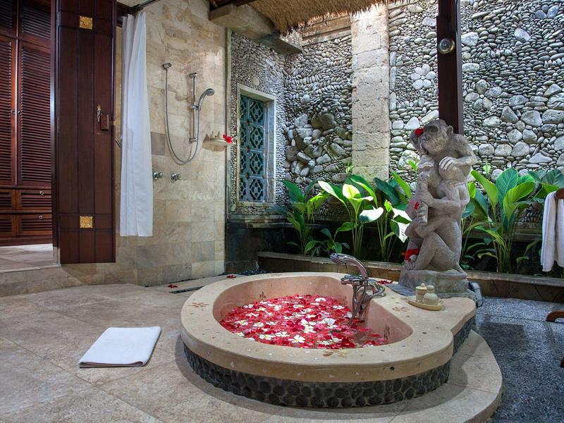 Luxusní vana s růžovými okvětními lístky v otevřené koupelně s kamennými zdmi a rostlinami.