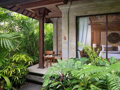 Open hotelkamer met bed, houten meubels en weelderige groene tropische begroeiing buiten.