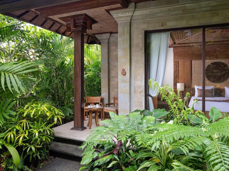 Open hotelkamer met bed, houten meubels en weelderige groene tropische begroeiing buiten.