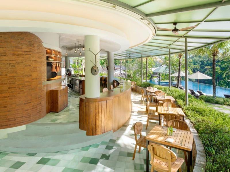 Espace piscine d'hôtel ouvert avec bar et plusieurs tables en bois à côté d'une piscine sous des parasols.