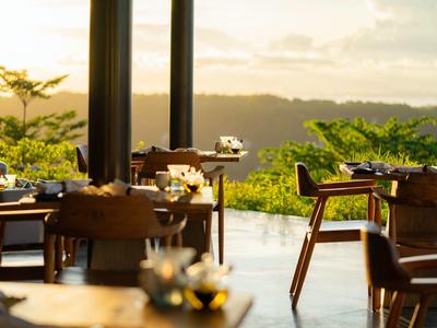 Restaurant avec tables et chaises en bois surplombant un paysage verdoyant au lever du soleil.