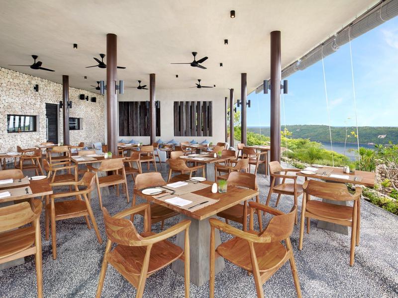 Espace repas ouvert avec chaises et tables en bois donnant sur un paysage vert et un ciel bleu.