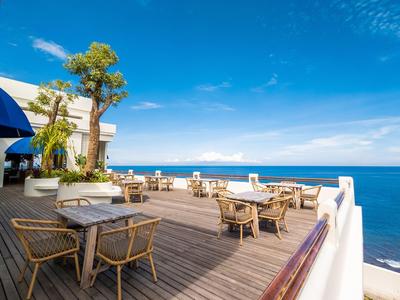 Freie Terrasse mit Holzstühlen und Tischen, blauer Himmel und Meer im Hintergrund.