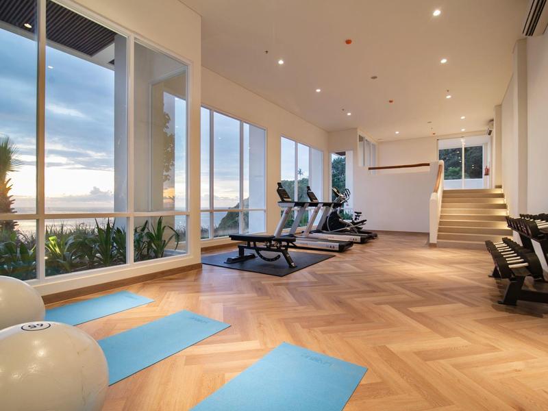 Heller Fitnessraum mit Holzfußboden, blauen Matten, Fitnessgeräten und großen Fenstern mit Meerblick.