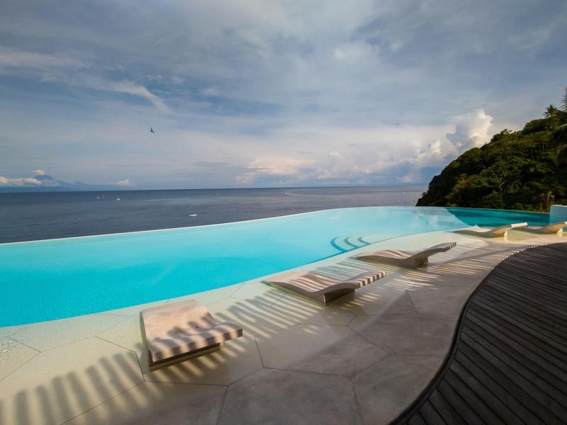 Infinity-Pool am Meer mit Sonnenliegen auf heller Terrasse und bewölktem Himmel.