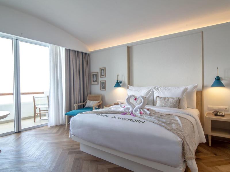 Helles Schlafzimmer mit großem Bett, Dekoration aus Handtüchern und Zugang zu Balkon mit Meerblick.