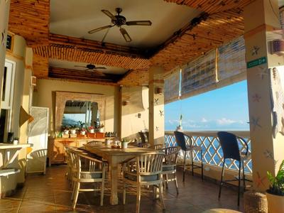 Terraza cubierta con mesas de madera, sillas y vista al océano bajo cielo despejado.