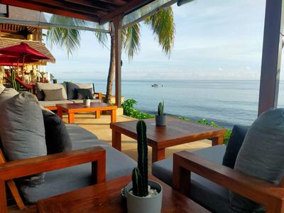 Offene Terrasse mit Holzmöbeln, grauen Kissen und Blick auf das Meer bei ruhigem Wetter.