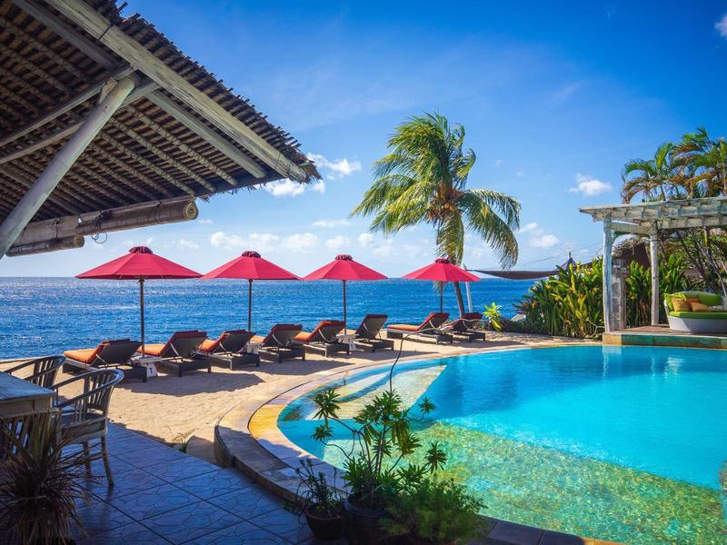 Zwembadgebied met uitzicht op de zee, ligstoelen en rode parasols onder een blauwe lucht.