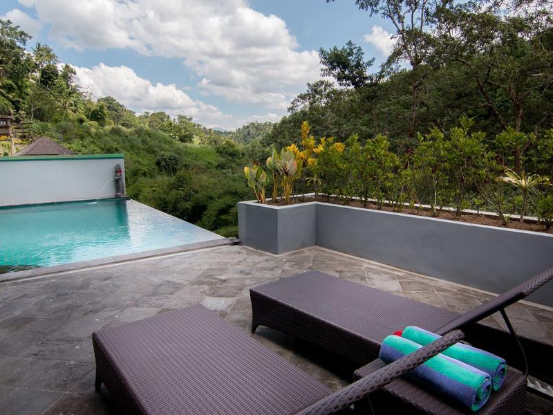 Taras z leżakami, krawędzią basenu i widokiem na zielone wzgórza pod niebieskim niebem.