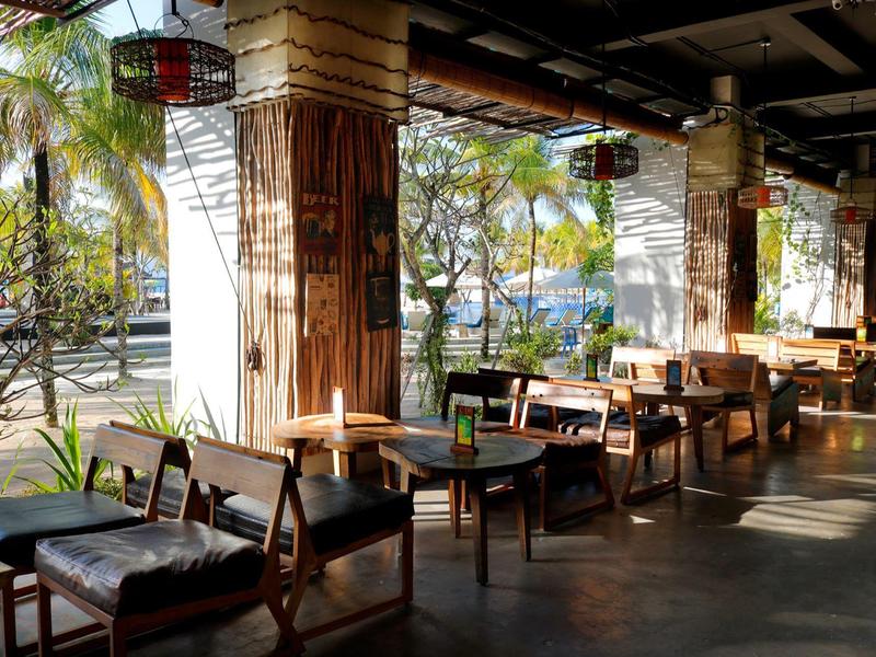 Terrazza soleggiata con mobili in legno e palme, vista su strada trafficata alla luce del giorno.